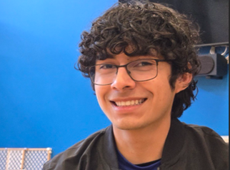 A person with curly hair and glasses smiles at the camera. They wear a dark jacket and a blue shirt, set against a bright blue background.