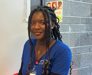 Young woman with long braided hair wearing a blue shirt, sitting by a brick wall. She is smiling softly, exuding a warm and friendly demeanor.