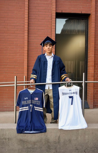 Young graduate in cap and gown holds two sports jerseys on hangers, one navy and one white, standing proudly against a brick wall background.