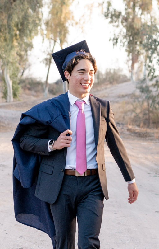 Young man in a suit with a pink tie and a graduation cap walks confidently outdoors, holding his gown, surrounded by trees and a sandy path.