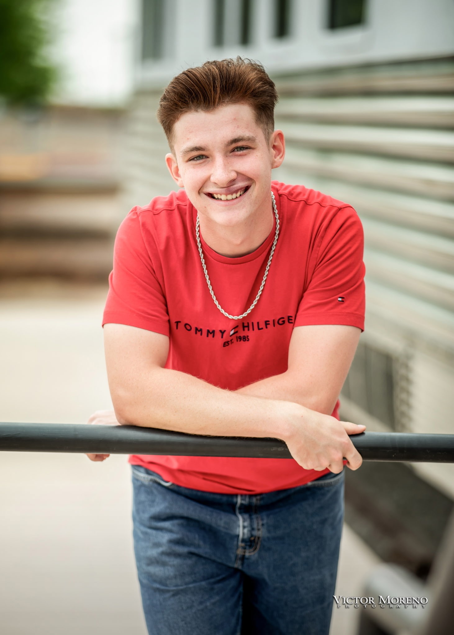 Young man smiling, wearing a red T-shirt and jeans, leans casually on a horizontal rail. Background features a blurred, modern building exterior.