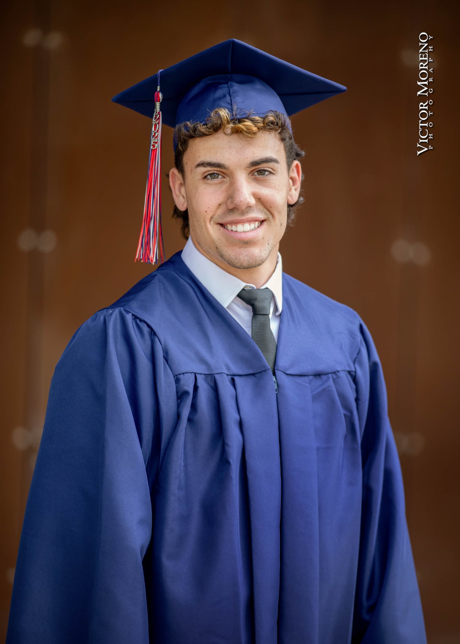 A young man smiles, wearing a blue graduation cap and gown with a red tassel, conveying pride and accomplishment against a blurred brown background.