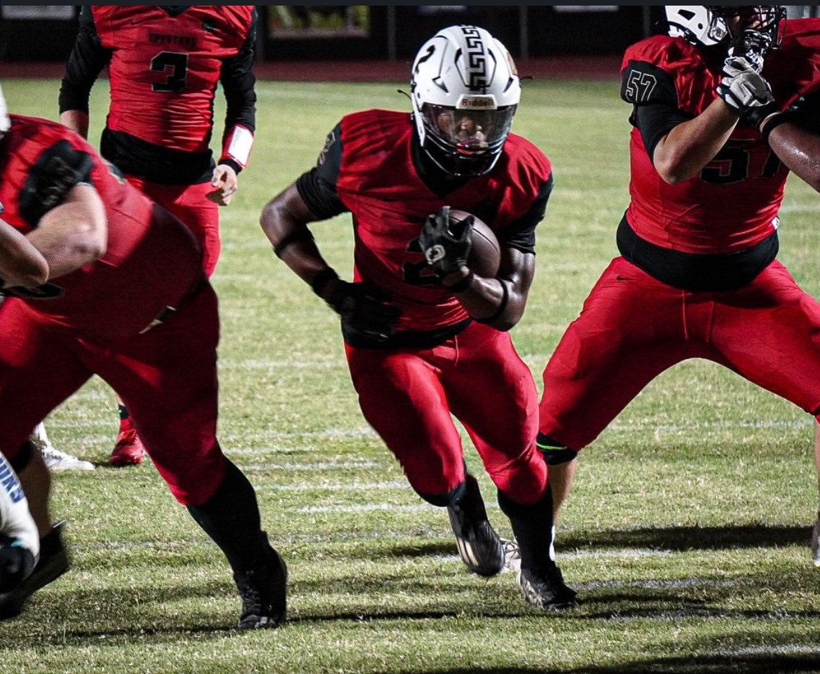 Football player in red uniform, helmet, running with ball, flanked by teammates. Night game setting. Intense focus and dynamic action.
