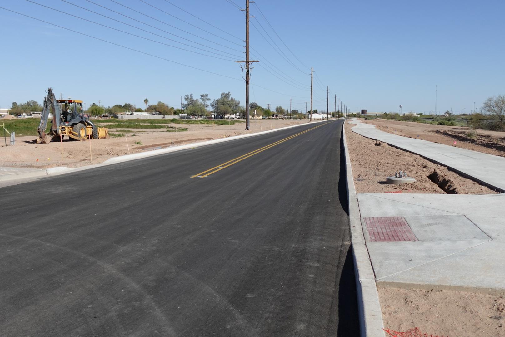 A newly paved road stretches into the distance under a clear blue sky. To the left, a digger sits idle on dirt, while sidewalks frame both sides of the road.