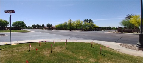 A wide, sunny suburban intersection with a green grass patch in the foreground marked by small red flags. Trees line the streets in the background.
