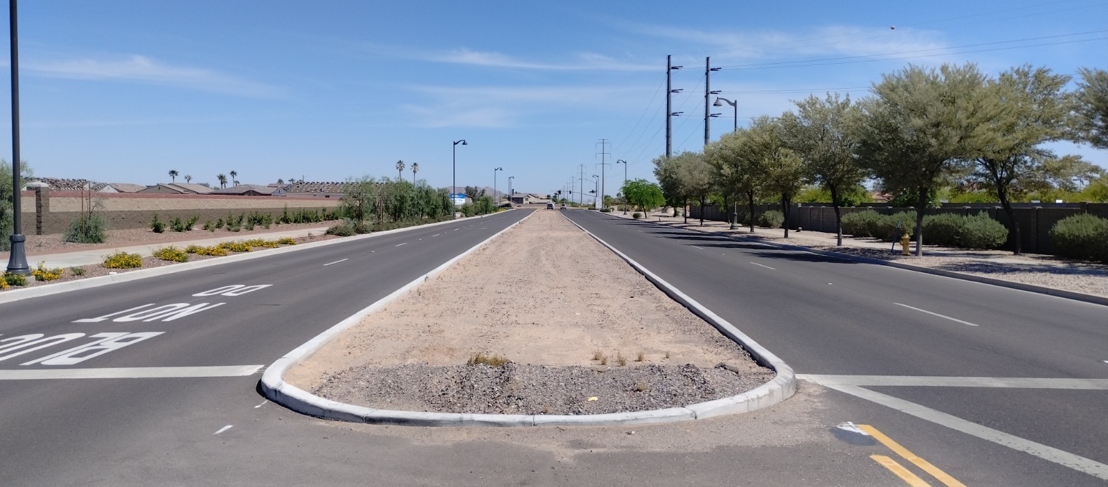 Wide, empty road with a dry, landscaped median, surrounded by low bushes and trees under a clear blue sky, suggesting a hot, sunny day.