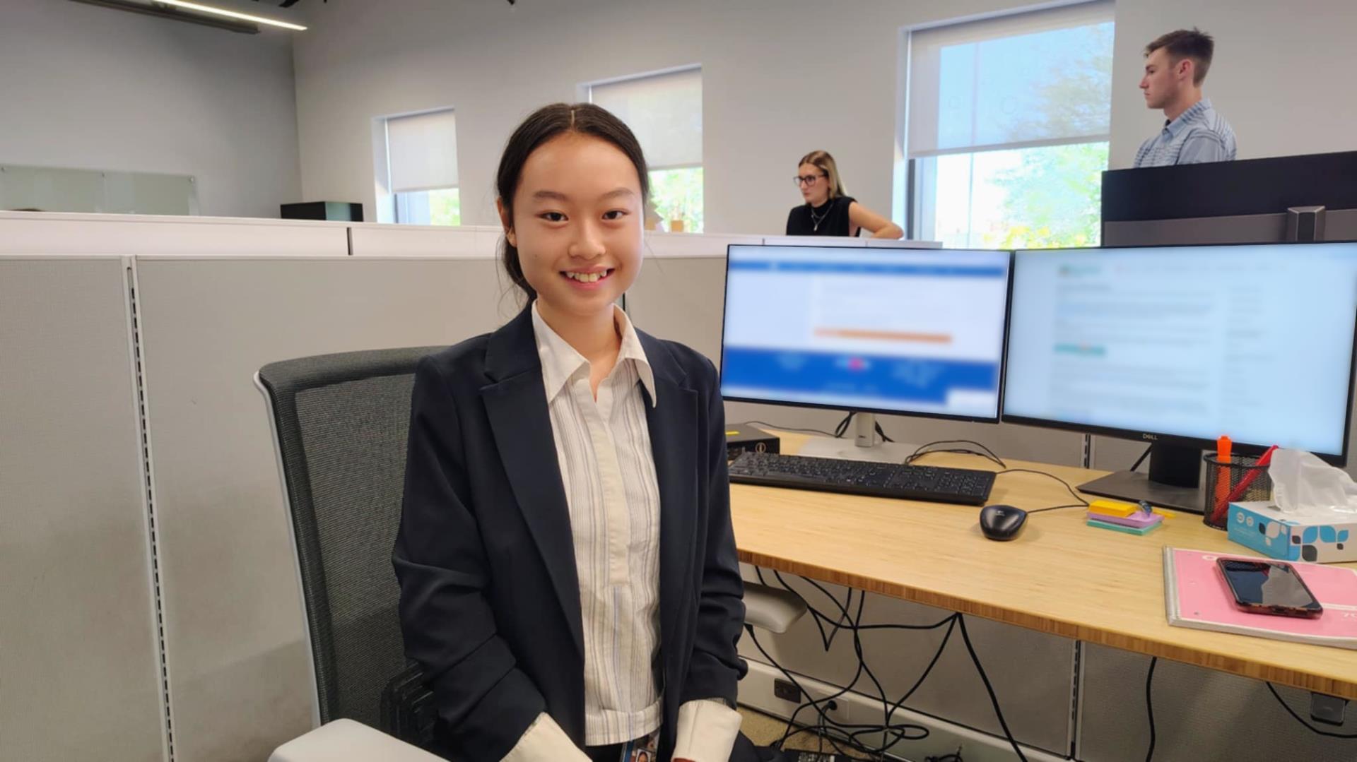 A young woman in a navy blazer smiles at her desk in an office. Behind her, coworkers are standing and working at computers, creating a professional and collaborative atmosphere.