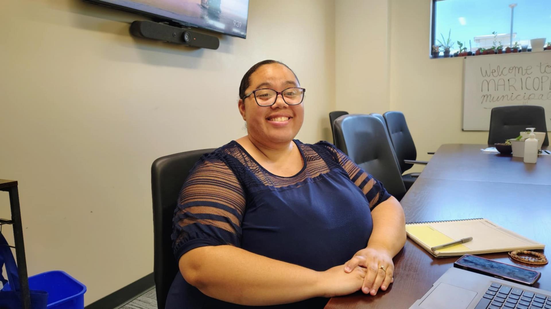 A smiling person sits at a conference table in an office, wearing a blue blouse. A notepad, phone, and laptop are on the table, with a welcoming sign in the background.