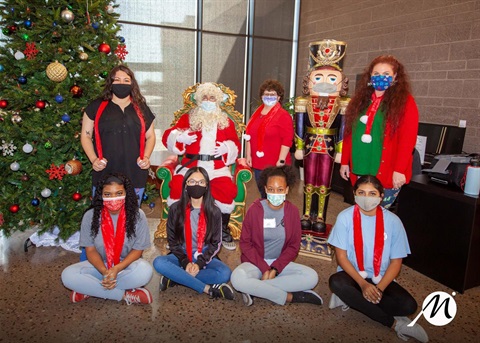 A festive holiday scene featuring masked individuals posing around Santa Claus beside a decorated Christmas tree and large nutcracker figure.