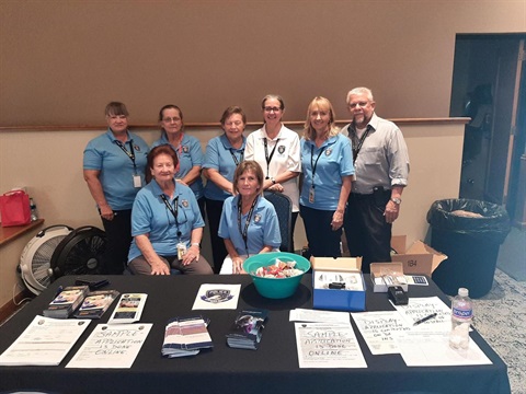 A group of eight people in matching blue shirts and ID badges pose behind a table with police-related brochures and forms. The mood is professional.