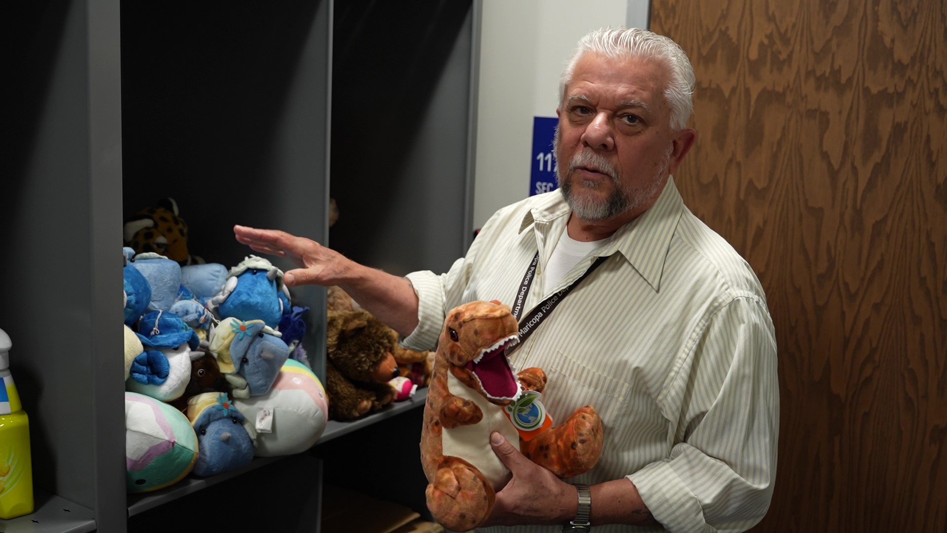 A man with white hair and a beard wearing a striped shirt stands beside shelves filled with colorful stuffed animals. He holds a plush dinosaur, looking thoughtfully at the camera.