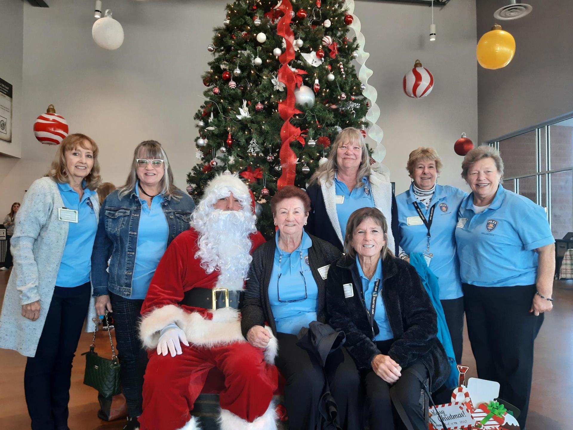 A group of eight people, including Santa Claus, pose cheerfully in front of a large Christmas tree decorated with red ribbons and ornaments.