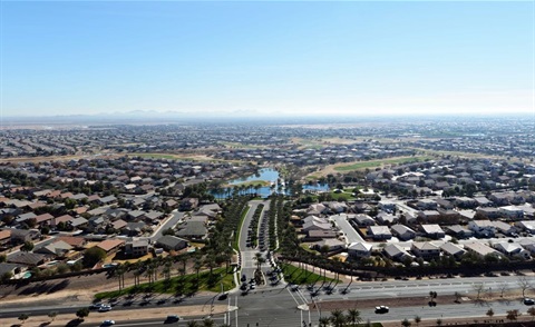 Aerial view of a sprawling suburban neighborhood with rows of houses, tree-lined streets, and small lakes. The sky is clear, giving a serene, expansive feel.