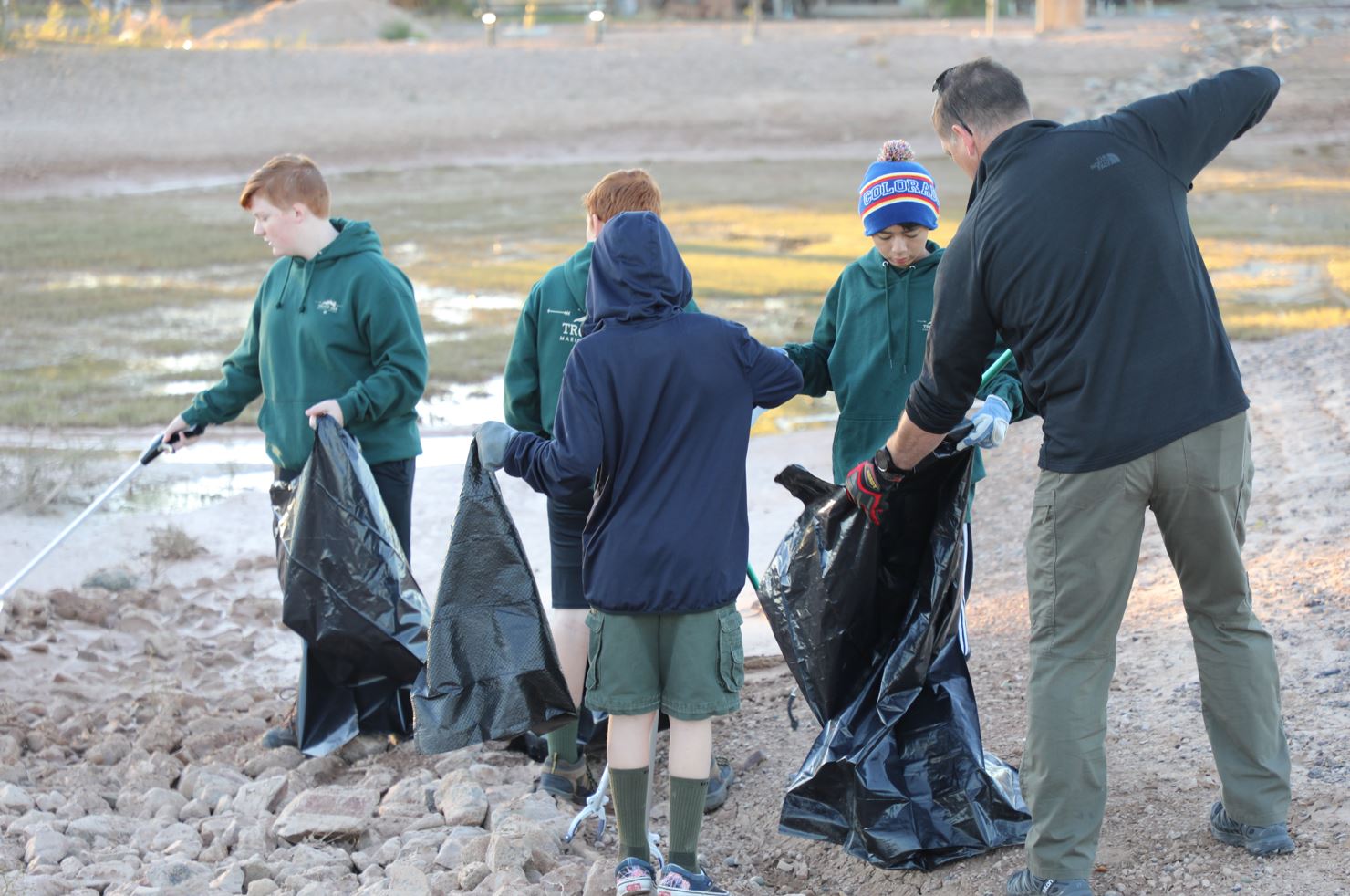 A group of young people and an adult in outdoor attire pick up litter with trash bags on a rocky, open area. The scene conveys teamwork and environmental care.