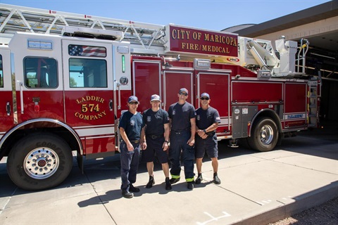 Four firefighters in uniform stand in front of a red fire truck marked 