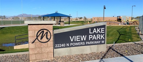 Sign for Lake View Park, featuring a logo and address, set against a backdrop of green lawns, a blue-roofed gazebo, and mountains under a clear sky.