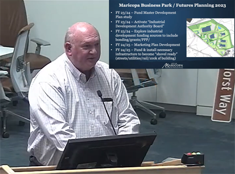 A bald man in a white shirt speaks at a podium in a conference room. Behind him, a slide titled 