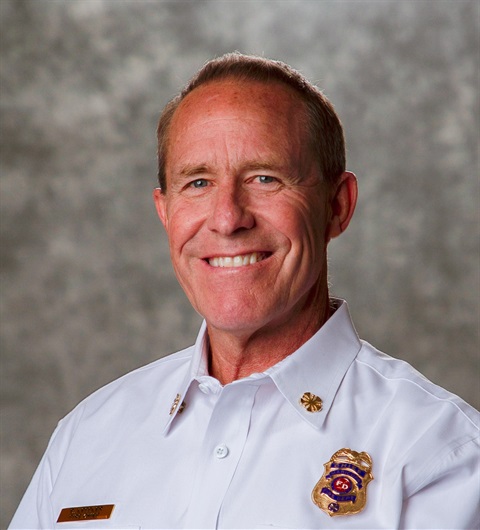 Smiling man in a white uniform with fire department badges, against a grey textured background. The image conveys professionalism and warmth.