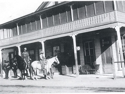 Vintage black and white photo of two people on horseback outside a wooden building with ornate railings and a sign reading 