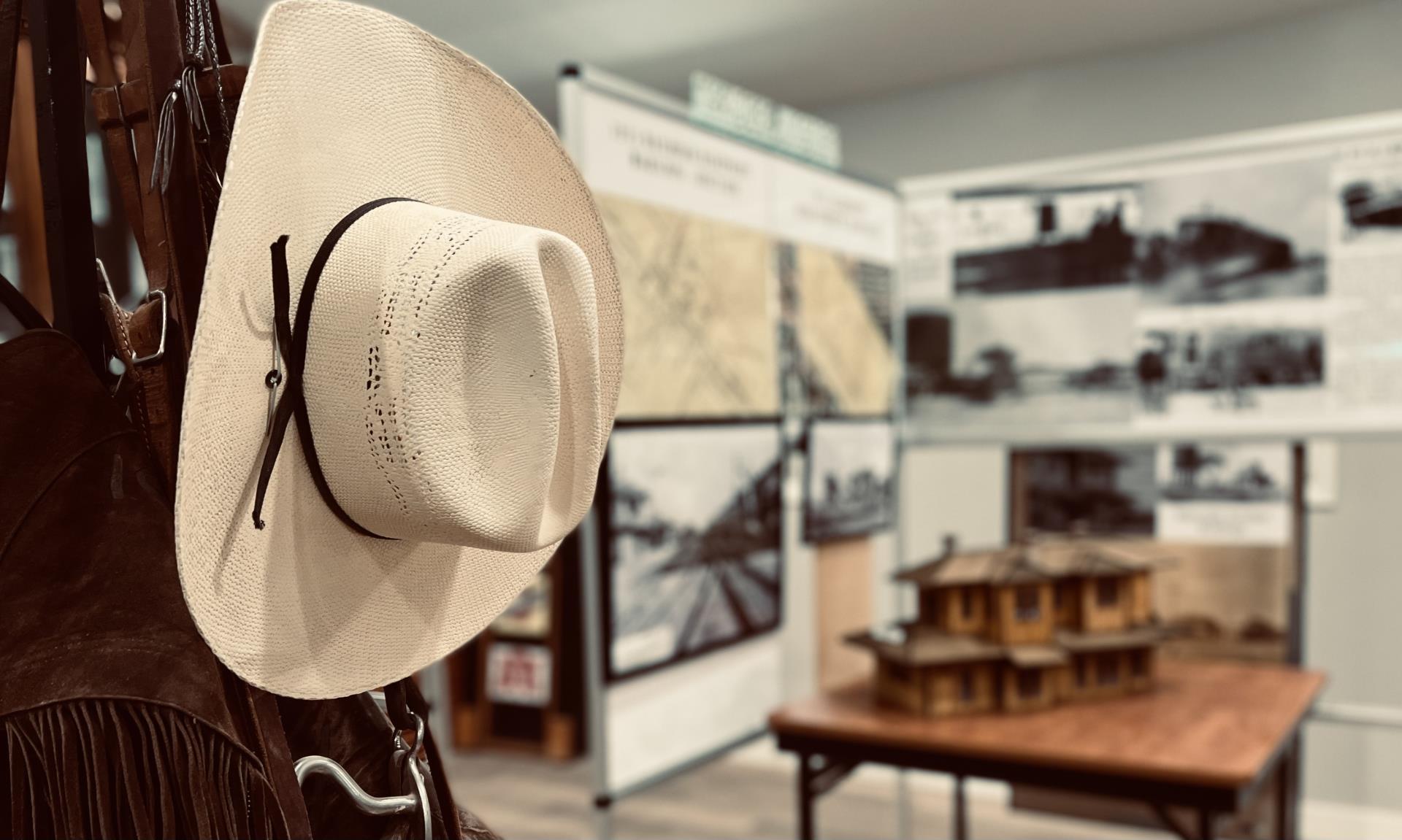 A white cowboy hat hangs with leather straps in the foreground, evoking a rustic feel. The blurred background shows a historical exhibit with maps and photos.