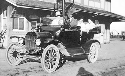 Vintage photograph of a classic early 20th-century car with three passengers and a driver in front of an old building. The scene conveys a nostalgic and historical atmosphere.