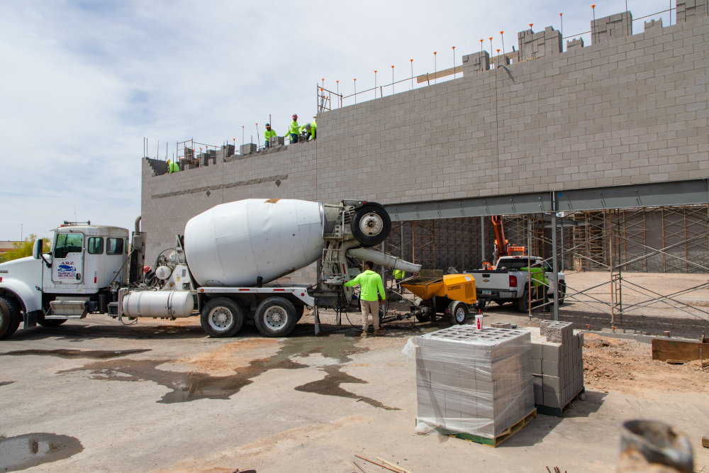 Construction scene with workers in green vests on a gray brick building. A cement truck unloads beside scaffolding; a pallet of bricks is in the foreground.