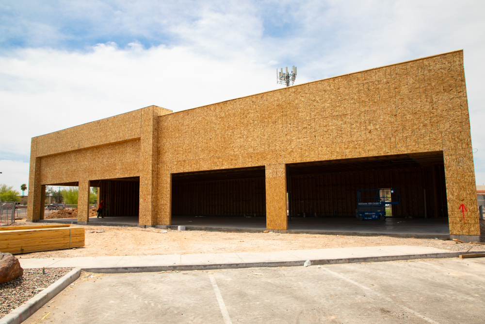 Unfinished commercial building with exposed plywood walls and open garage-like spaces. Construction materials are nearby under a clear sky.