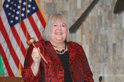 A woman in a red and black patterned jacket smiles while holding a gavel. An American flag is visible in the background. The setting appears formal.