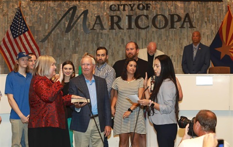 A woman in a red jacket takes an oath with a hand on a book, surrounded by people on a stage. U.S. and Arizona flags are displayed. The mood is formal.