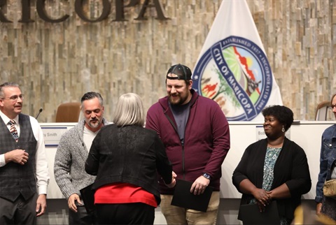 A group of people stands in an indoor setting, some holding certificates. A woman shakes hands with a man in a cap. A city emblem is visible in the background.