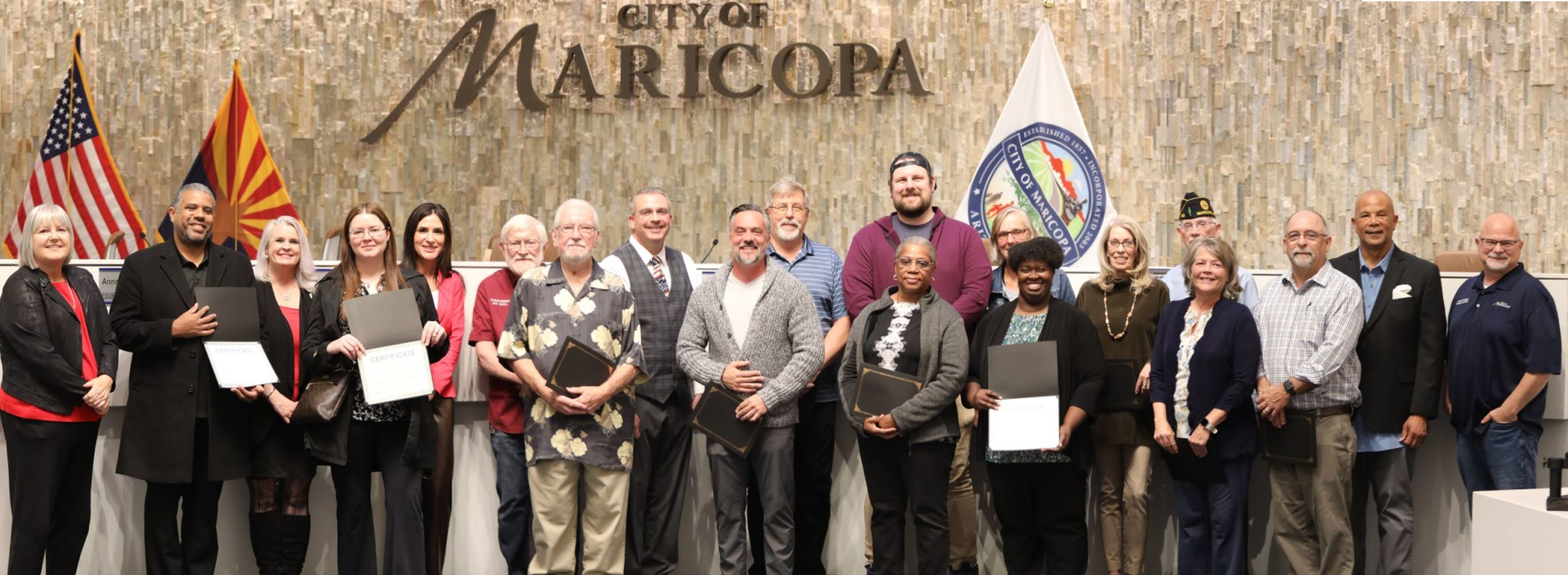 A diverse group of smiling people holds certificates while posing in front of a 