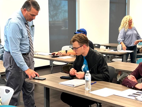 A man in a tie shows a cellphone to a seated student with glasses in a classroom. Papers and a water bottle are on the desk. Others converse in the background.