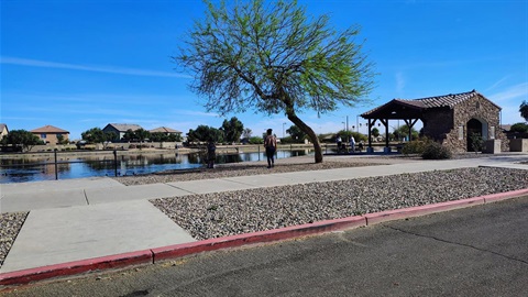 A person walks on a paved path beside a tranquil pond, with a single tree and a rustic stone pavilion under a clear blue sky, conveying serenity.