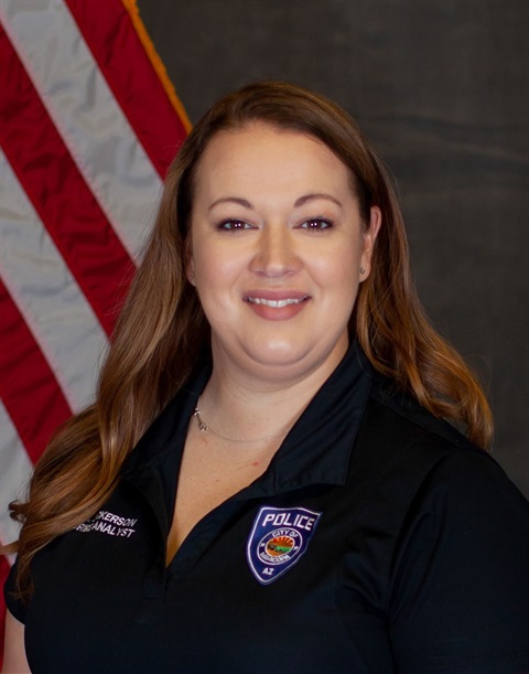 Smiling woman in a black police uniform shirt stands in front of a U.S. flag. The setting conveys professionalism and pride.