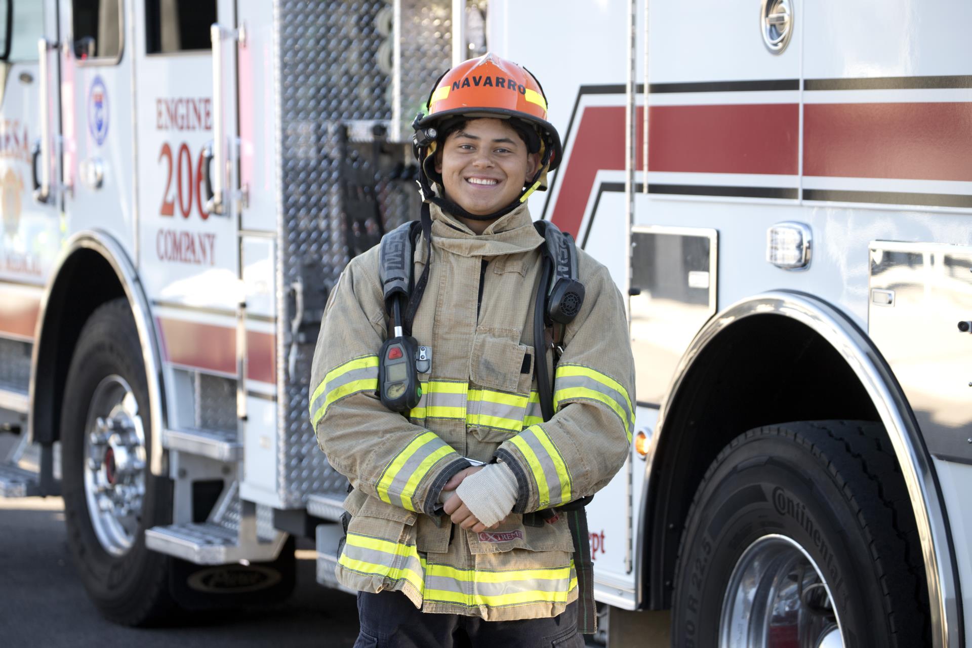 Smiling firefighter in full gear stands beside a fire truck with reflective stripes, conveying confidence and readiness. Daytime setting.