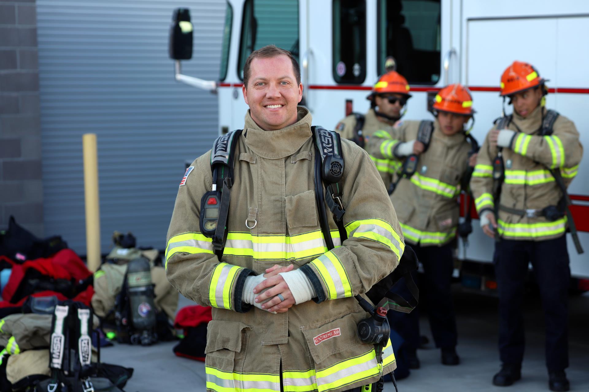 Smiling firefighter in full gear stands in front of a fire truck. Three colleagues in helmets prepare in the background. Scene conveys readiness and teamwork.