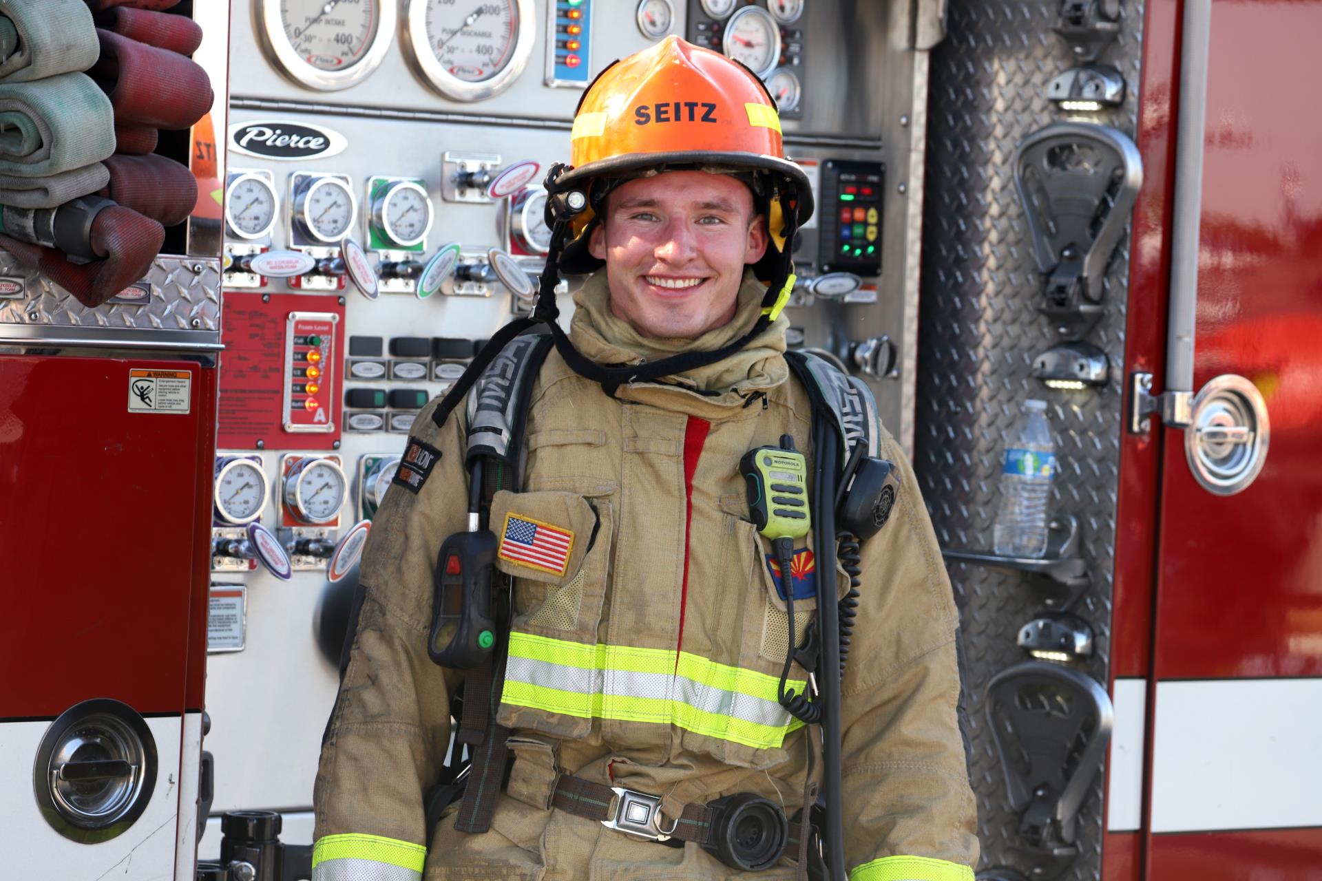 A firefighter in gear stands smiling in front of a fire truck panel, with gauges and equipment, conveying readiness and dedication.