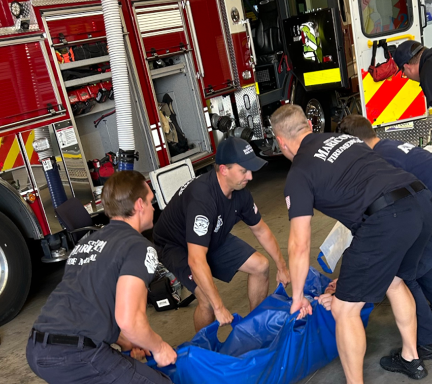 Four firefighters are practicing lifting techniques with a blue rescue tarp in a station, red fire truck in the background, showing teamwork and focus.