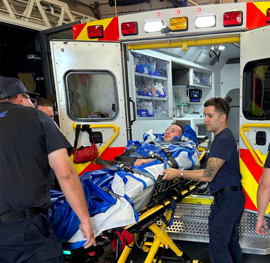 Paramedics load a patient secured to a stretcher into an ambulance. The vehicle's interior is visible, conveying urgency and medical care.