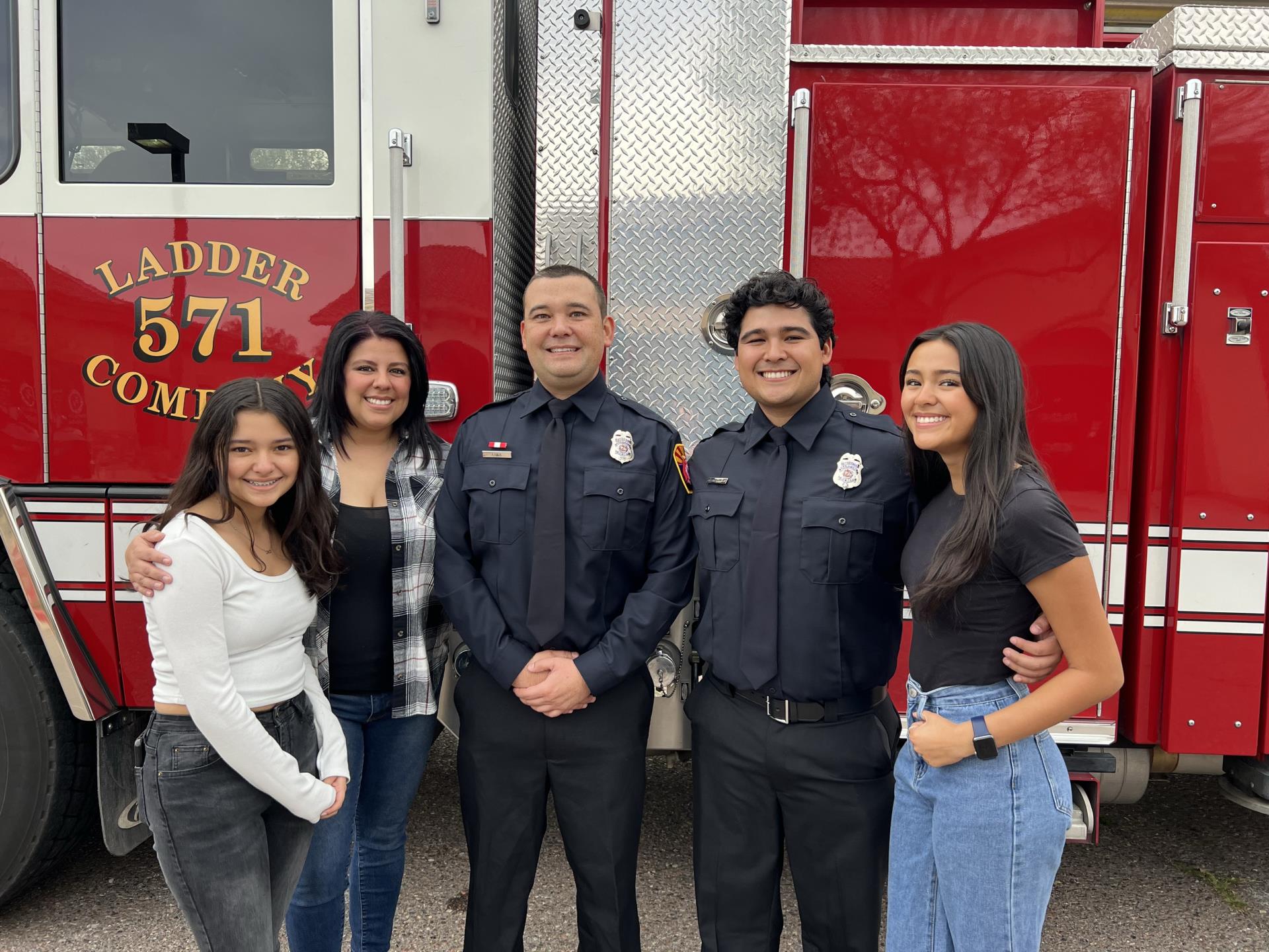 A group of five people, including two in firefighter uniforms, stand smiling in front of a red fire truck labeled 