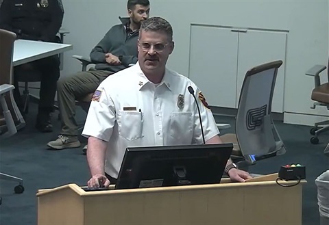 A man in a fire department uniform speaks at a podium in a conference room. He appears focused, surrounded by seated attendees in casual attire.