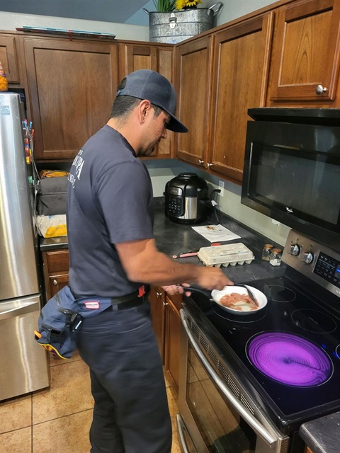A man in a cap cooks on a stove in a kitchen. He is holding a pan with food. Wooden cabinets, a fridge, an egg carton, and a pressure cooker are visible.