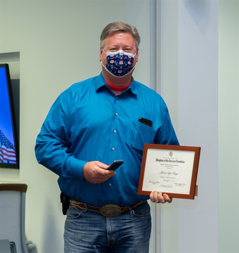 Man in a bright blue shirt and jeans holds a framed certificate and a phone. He wears a patterned mask, standing in a room with neutral walls.