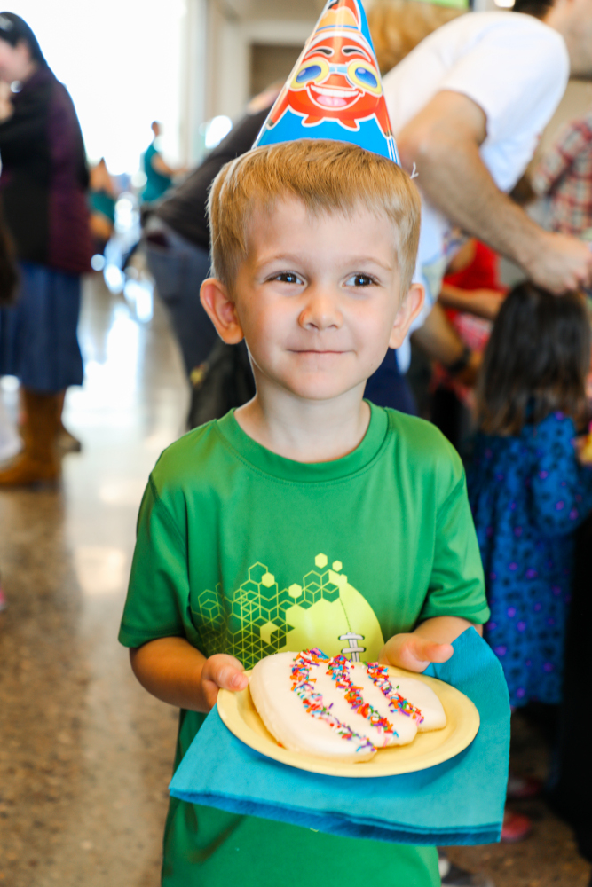 Young child wearing a festive party hat and green shirt holds a plate with a frosted cookie topped with sprinkles, smiling amidst a joyful gathering.