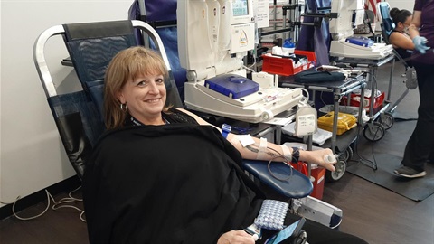 A woman, smiling and relaxed, donates blood while seated in a donation chair. Medical equipment and a healthcare worker are visible in the background.