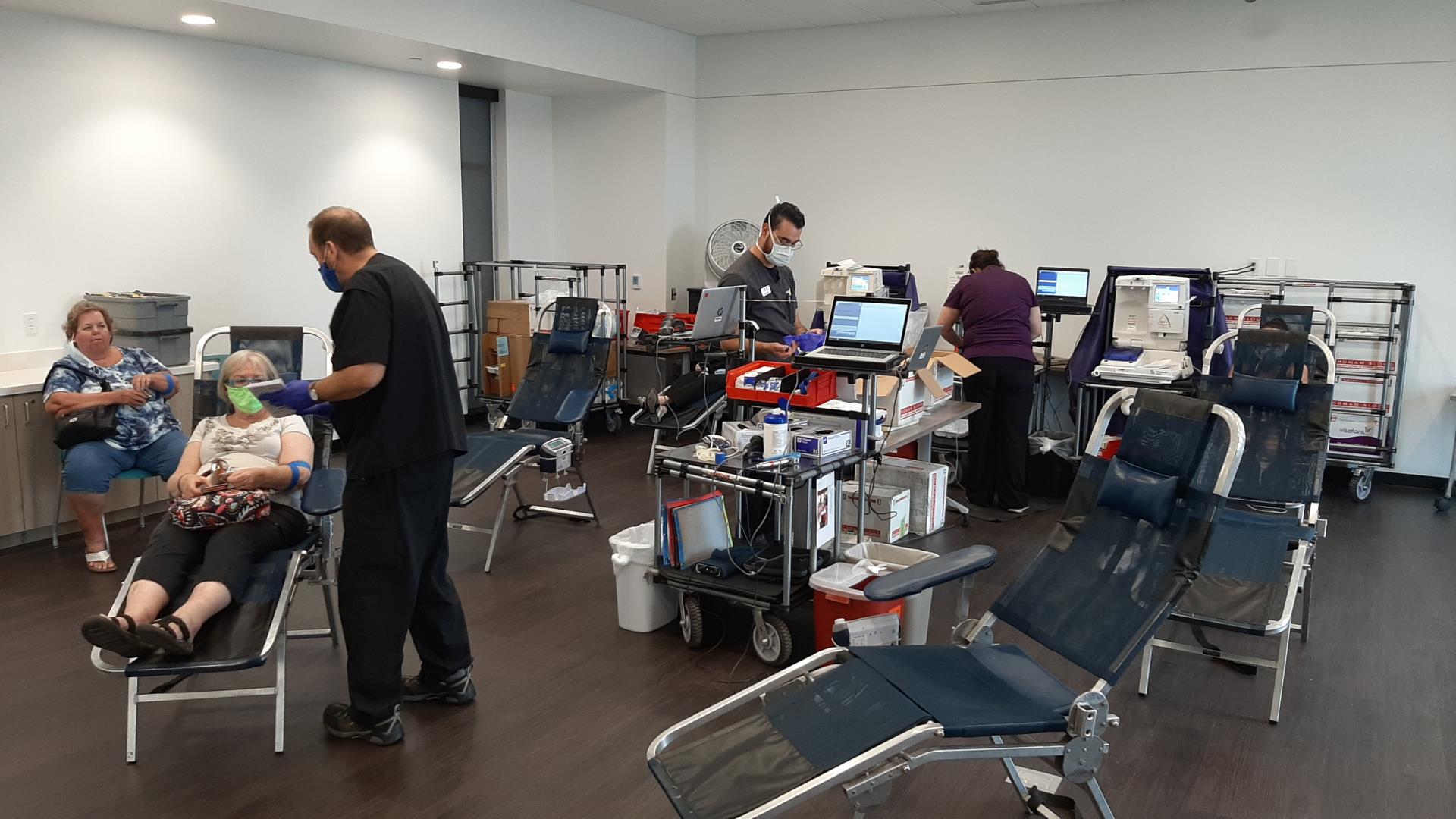 A busy blood donation center with four people. Donors lie on chairs, staff assist, and medical equipment is organized on carts. Calm, clinical atmosphere.