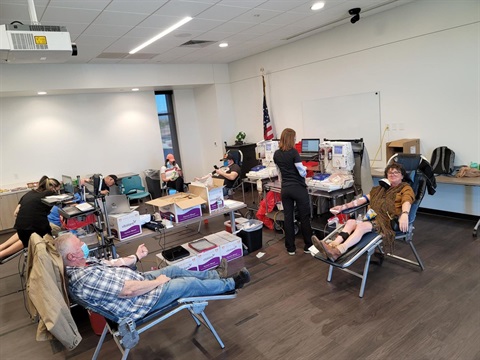 Room with people donating blood, lying on chairs with medical equipment nearby. A staff member oversees the process. A U.S. flag is in the background.
