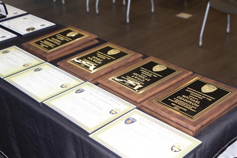 A table displays several award plaques and certificates on a black cloth. The wood plaques have golden engravings, creating a formal and celebratory tone.