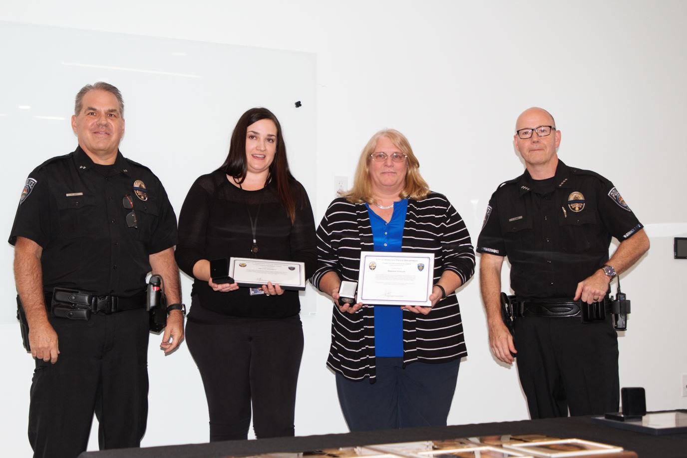 Four people stand in a row, two in police uniforms and two women holding certificates, smiling, in a bright room, conveying a sense of achievement.