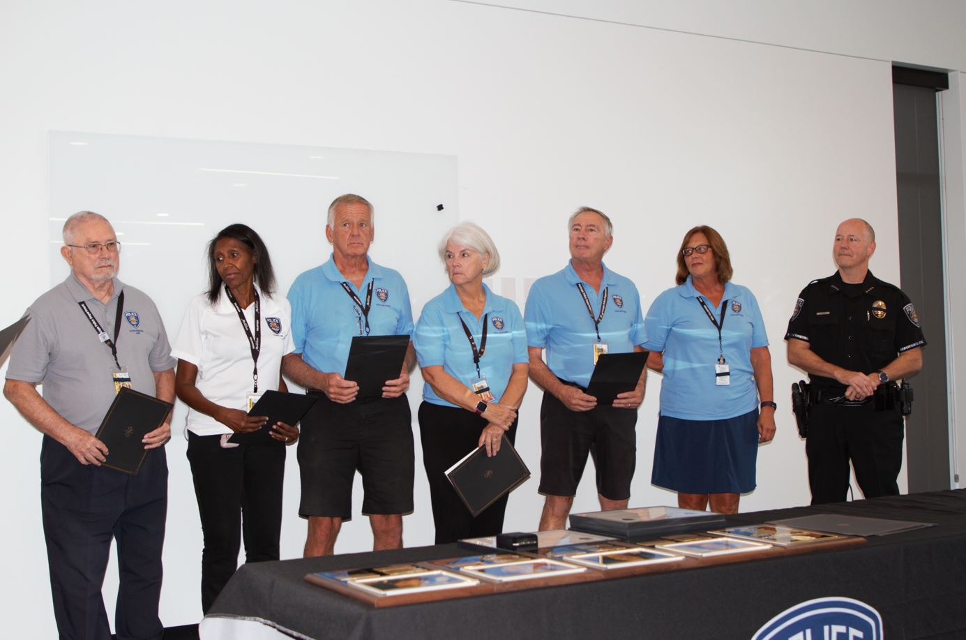 A group of six people stands in front of a whiteboard, five in light blue shirts holding certificates. A police officer is on the right. The setting is formal.